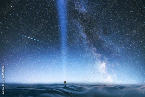 Fototapeta Naklejka Na Ścianę i Meble -  Fantasy night landscpe. The person with flash light stands on the hill covered long exposhur clouds. Behind the hill bright milky way galaxy on the starry sky. 