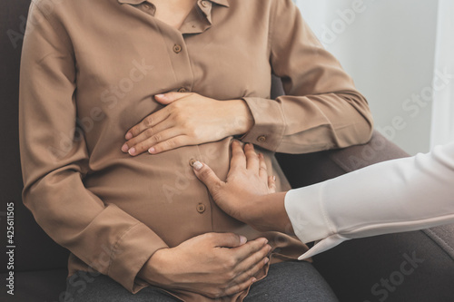 Young asian pregnant woman holding her belly while gynecologist check up the symptoms by touching her abdomen about the pregnancy is explaining about the unborn child.