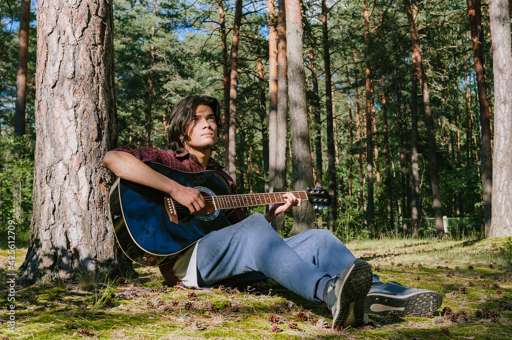 The guy plays the guitar sitting near the tree. Against the background of the forest.