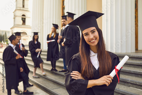 Portrait of a happy smiling graduate girl dressed in a black mantle with a diploma in her hands on the background of her classmates. Student poses on the steps of the university. photo