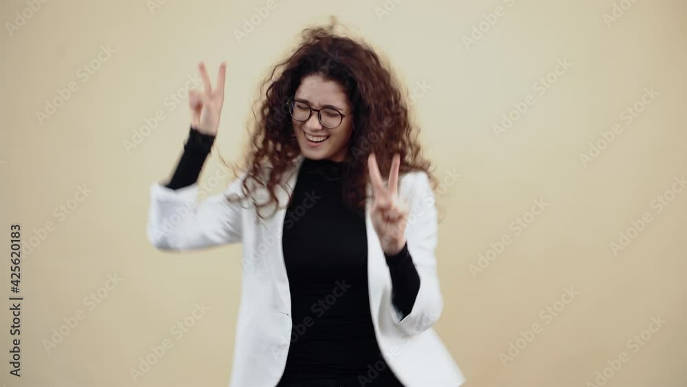 The distracted young woman with curly hair dances with her fingers in the shape of a peace sign, shaking her hands. Young hipster in gray jacket and white shirt, with glasses posing isolated on beige The distracted young woman with curly hair dances with her fingers in the shape of a peace sign, shaking her hands. Young hipster in gray jacket and white shirt, with glasses posing isolated on beige