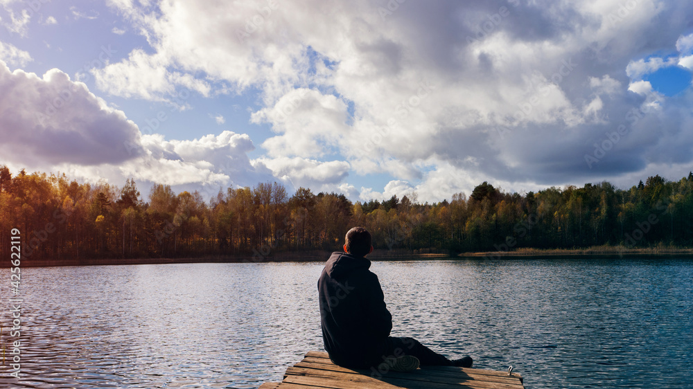 alone young man sits on a wooden bridge and enjoys the warm autumn and beautiful scenery on the lake shore