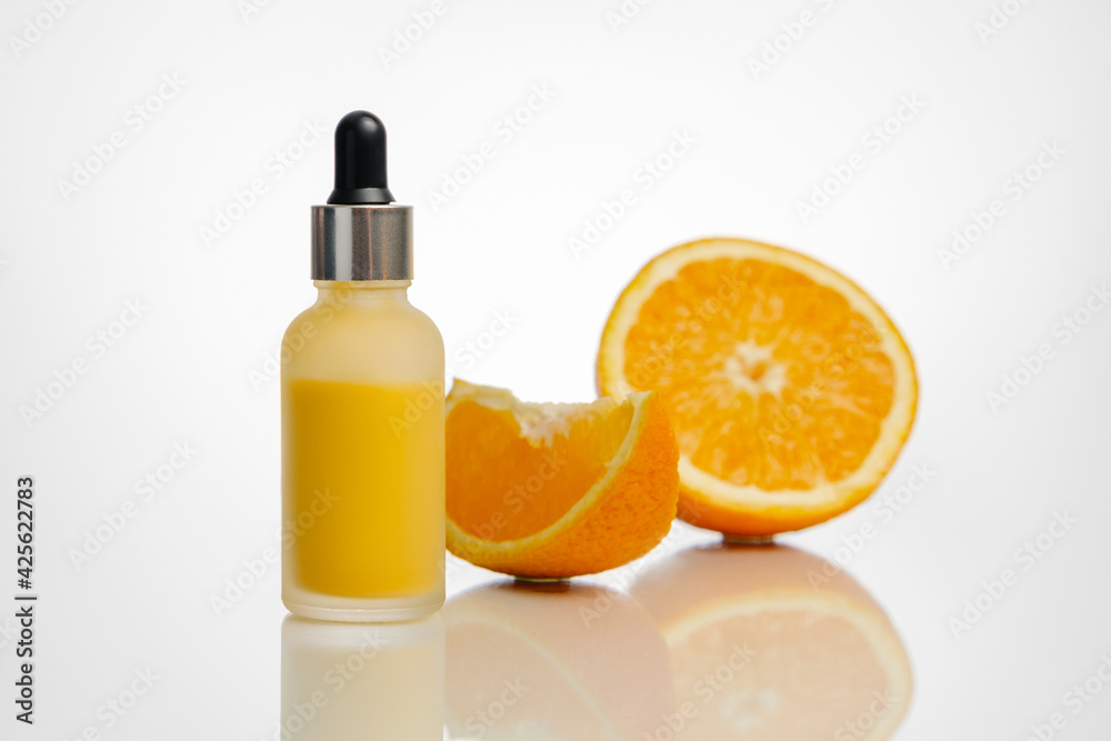 A bottle of orange serum and some oranges in the background.