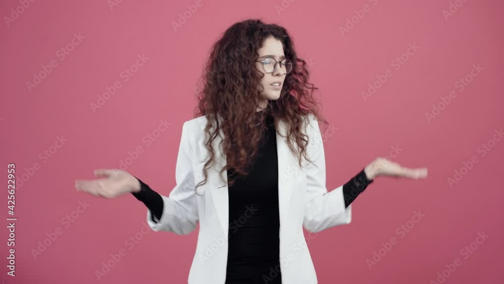 The young woman is confused with her curly hair, gestures with her hands and does not understand why, she had moved from side to side. Young hipster in white jacket and black shirt, with glasses The young woman is confused with her curly hair, gestures with her hands and does not understand why, she had moved from side to side. Young hipster in white jacket and black shirt, with glasses