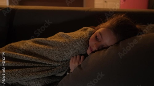 A cute girl falls asleep in front of the TV on the sofa, covered with a blanket.