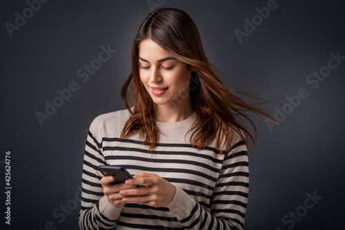 Smiling young woman text messaging while standing at isolated dark background photo