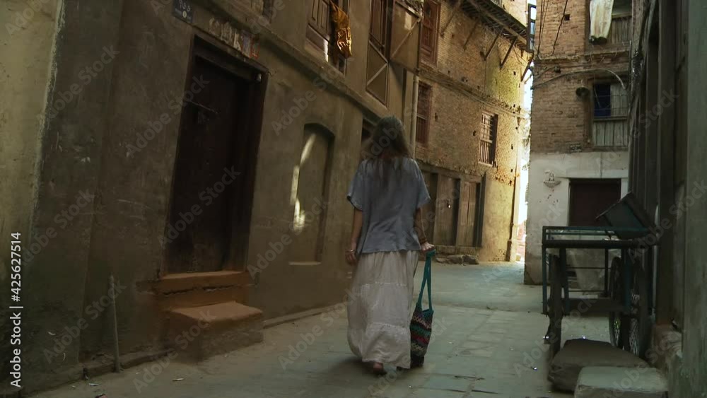 View fro behind of a long haired hippie girl with long white gown strolling in the roads and narrow alleys of Katmandu. Touristic activity in Nepal.