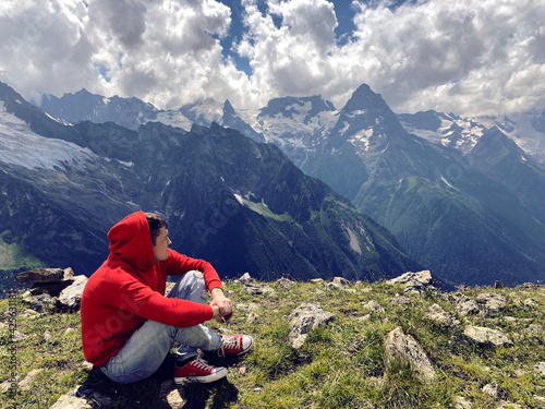 Young man sitting on mountain peak in cloudy weather. Adult guy rests, enjoying amazing mountain landscape in summertime.