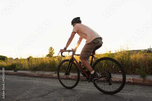 Young sporty guy in a helmet rides on gravel bicycle on asphalt road at sunset.