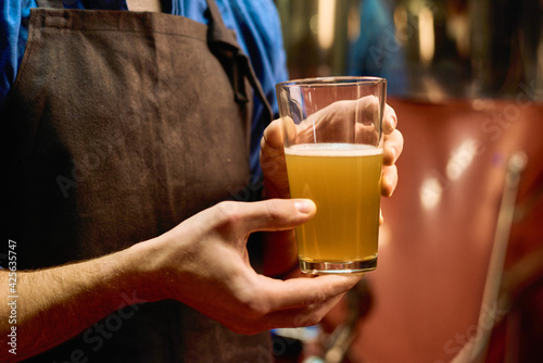 Hands of young male brewer in apron holding glass of fresh beer after preparation while standing in front of camera in workshop photo