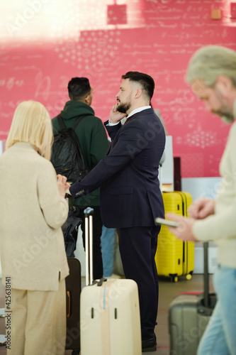 Contemporary businessman in elegant suit talking on mobile phone while standing in queue for registration before flight inside airport