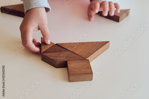 Close up kid's hand holding a piece of an wooden tangram puzzle on white table. Thinking. Mental developing. Playing, fun