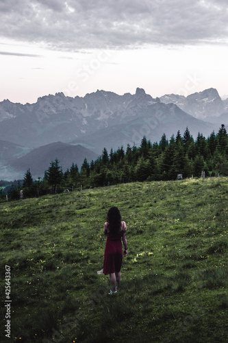 Back view of slim young woman standing on grassy valley on background of green mountains on cloudy evening