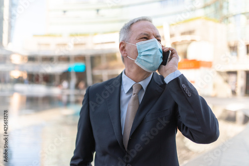 Covid coronavirus concept, masked elder man talking on his smartphone outdoor