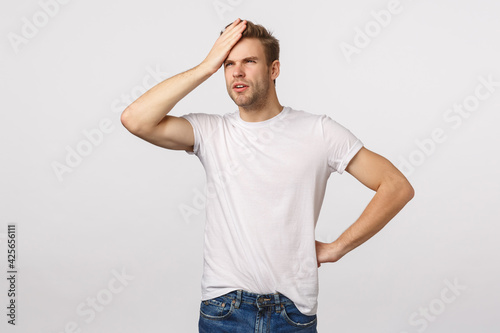 Focused and thoughtful young handsome blond bearded male in white t-shirt, punch forehead, squinting and looking upper left corner at sun, feeling head, checking tempreture or being forgetful photo