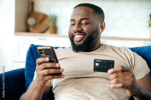 Online shopping. Happy confident young african american man in casual clothes sits on the couch at home, holds smartphone and banking card in his hands, pays for online purchases, entered card number photo
