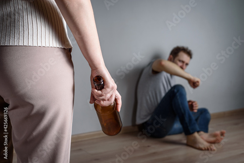 A drunk woman with an empty alcohol bottle in her hand stands over her frightened husband sitting on the floor. Female alcoholism and domestic violence.
