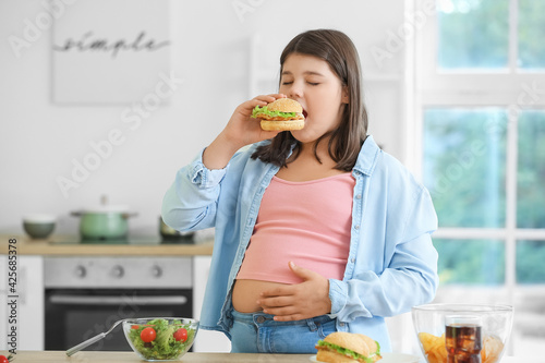 Fotografie Overweight girl eating unhealthy burger in kitchen