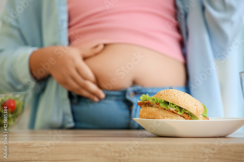 Plate with unhealthy burger on table near overweight girl photo