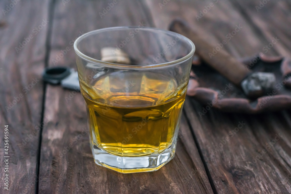 A glass of whiskey stands on a wooden table. Against the background of a cigar with an ashtray out of focus A glass of whiskey stands on a wooden table. Against the background of a cigar with an ashtray out of focus