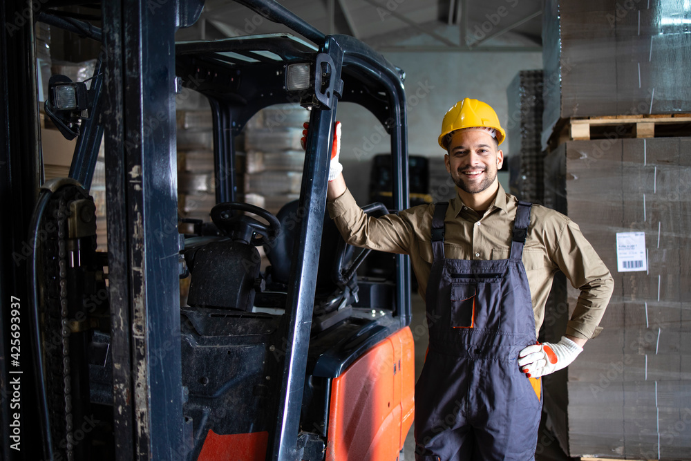 Portrait of experienced young caucasian forklift driver standing by loading cargo machine in storage warehouse. Portrait of experienced young caucasian forklift driver standing by loading cargo machine in storage warehouse.