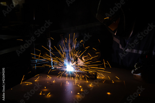 A man welder with construction gloves and a welding mask is welded with a welding machine metal in workshop.