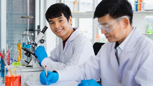 Asian young man sitting work with microscope and adult man wearing eyeglass do writing a note book both are chemist scientist and in uniform.Test tube with liquid put on table in laboratory