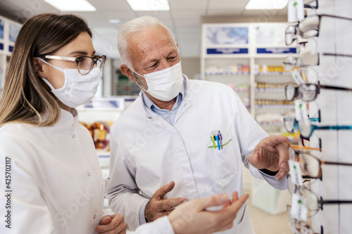 Conversation of two pharmacists about diopter for glasses. Woman with glasses and an older man stack glasses in a pharmacy. They wear white uniforms and masks to protect themselves from the virus