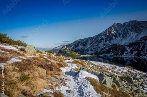 Blue sky over High Tatra Mountains in winter. Withered grass, cold lake in the valley and mountain range in the shadow. Selective focus on the rocks, blurred background.