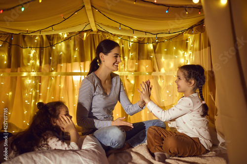 Family leisure time, motherhood and childhood concept. Happy loving mother playing with two adorable daughters in home tent decorated night lights lamp in evening before bedtime