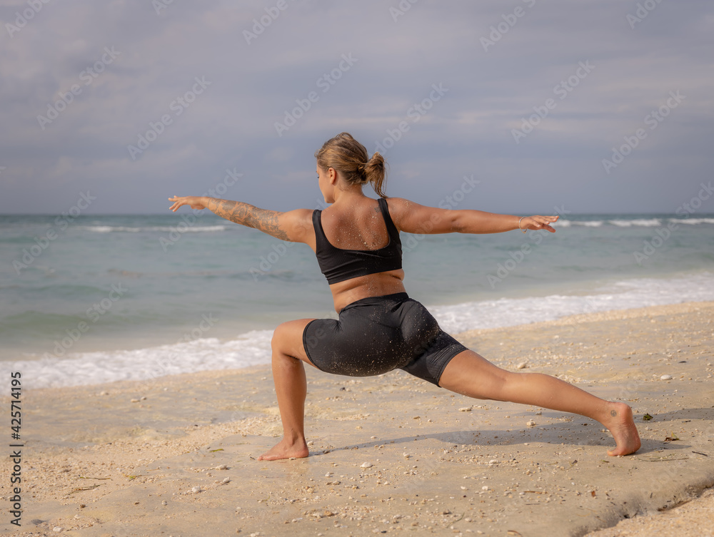 Young woman practicing yoga. Standing in Virabhadrasana II pose, Warrior II Pose. Work out. Healthy lifestyle. Strong body. Ocean background. Yoga retreat. Thomas beach, Bali