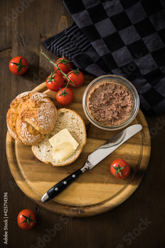 Grobe Leberwurst im Glas aus Vorrat mit Brötchen, Butter und Tomaten als Vesper Brotzeit auf Brotbrett und Holz Hintergrund dunkel und rustikal