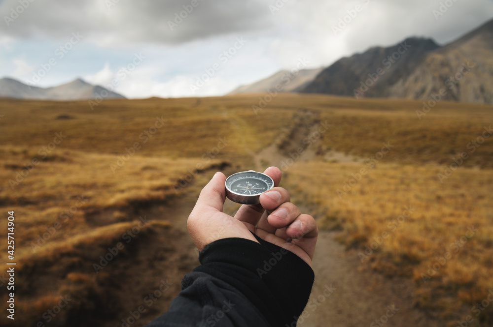 Man's hand holding a magnetic compass first-person view against the background of a high-altitude path and mountains - obrazy, fototapety, plakaty 