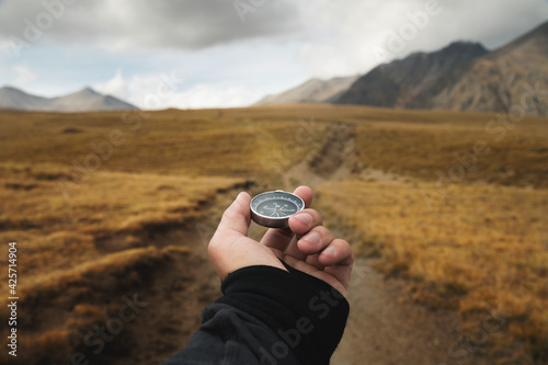 Man's hand holding a magnetic compass first-person view against the background of a high-altitude path and mountains photo