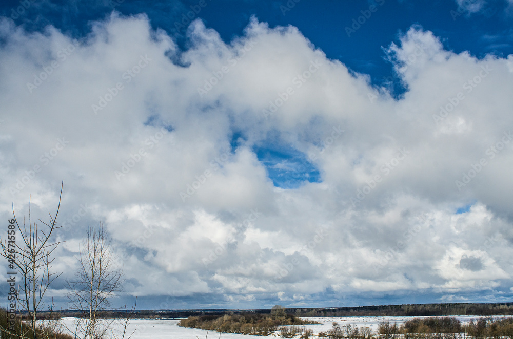 Winter landscape with a high bank of the river covered with ice, cumulus clouds in the sky,.deep snow cover with footprints, a clear horizon line.