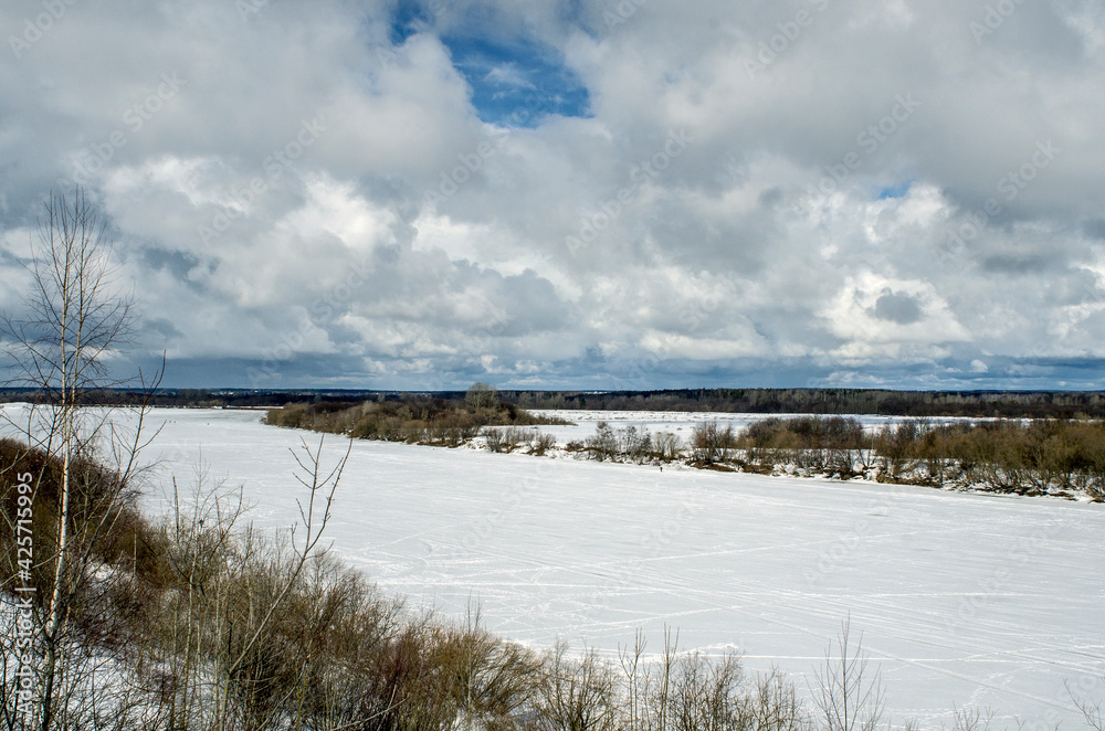 Winter landscape with a high bank of the river covered with ice, cumulus clouds in the sky,.deep snow cover with footprints, a clear horizon line.