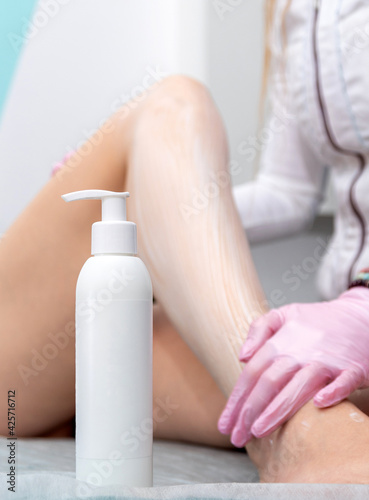 A white bottle of cream for advertising. Against the background of a care procedure, a gloved hand smears a woman's foot with cream. Selective focus.