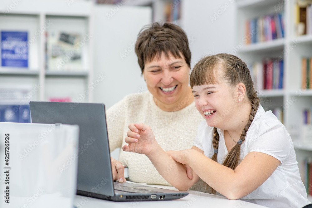 Smiling girl with down syndrome is uses a laptop with her teacher at library. Education for disabled children concept Smiling girl with down syndrome is uses a laptop with her teacher at library. Education for disabled children concept