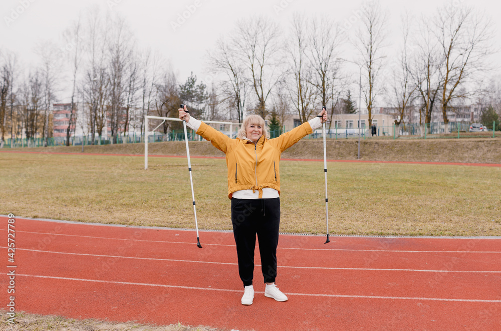 An elderly woman in a yellow sports jacket does physical exercises with Nordic sticks in her hands at the stadium.