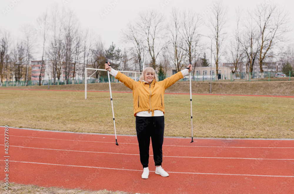 An elderly woman in a yellow sports jacket does physical exercises with Nordic sticks in her hands at the stadium. An elderly woman in a yellow sports jacket does physical exercises with Nordic sticks in her hands at the stadium.