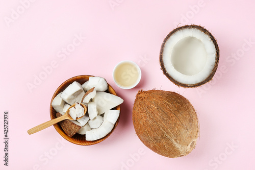 Coconut as a food source and cosmetic product. Cracked fruit in a wooden bowl with a jar of moisturizing oil on pink tabletop. Close up, top view, copy space, flat lay, background.
