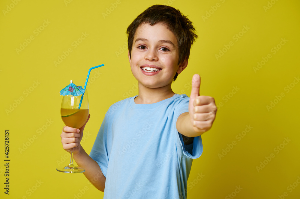 Happy boy holding glass with delicious fruit drink, decorated with cocktail umbrella and showing thumb up, smiling cute at camera on yellow background with copy space
