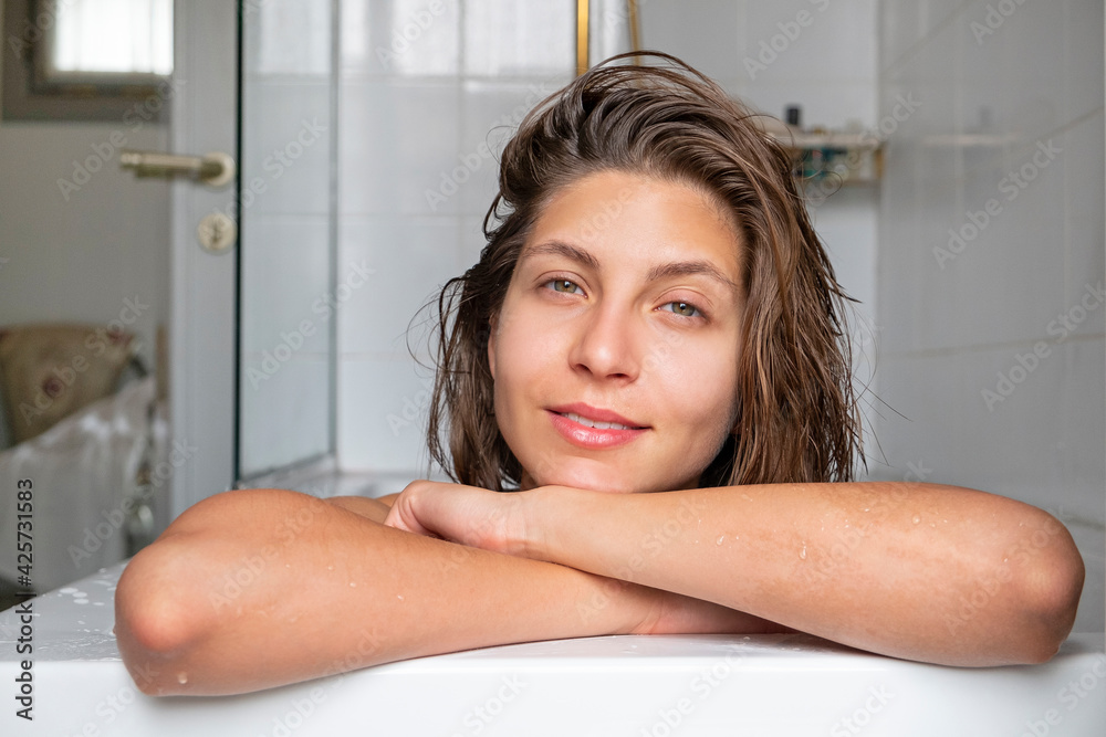 Obraz premium Close up portrait of a beautiful smiling young woman relaxing in a bathtub. Happy brunette female chilling in a bath of hotel room. Copy space, background.