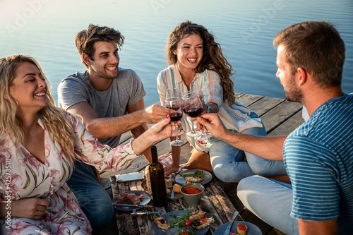 Friends having fun on picnic near a lake, sitting on pier eating and drinking wine. © Zoran Zeremski