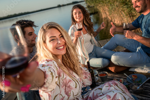 Friends having fun on picnic near a lake, sitting on pier eating and drinking wine. © Zoran Zeremski