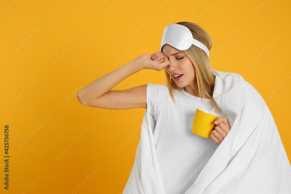 Young woman in sleeping mask wrapped with blanket holding cup on yellow background