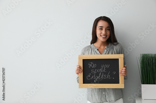 Young female teacher holding chalkboard with words DO YOU SPEAK ENGLISH? on light background. Space for text
