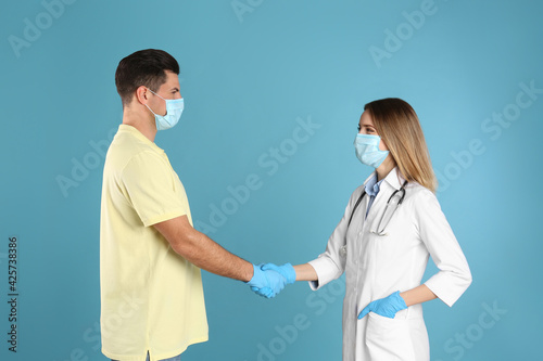 Doctor and patient in protective masks shaking hands on light blue background