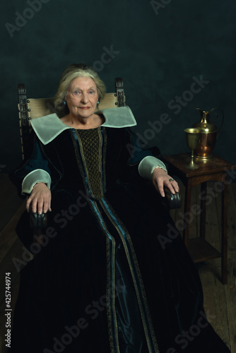 Canvas Print Senior woman dressed in renaissance attire sitting in a chair by a small table topped with a copper pitcher and a tin goblet near a dark wall