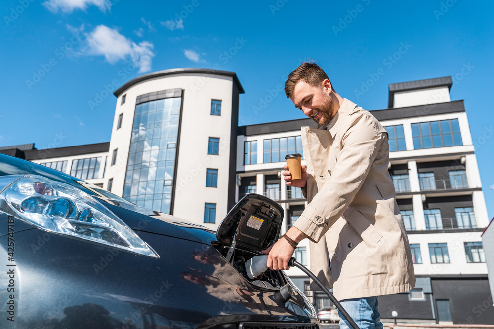 Man with coffee cup in hand inserts plug into the charging socket of electric mobile Man with coffee cup in hand inserts plug into the charging socket of electric mobile